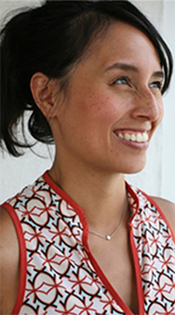 A smiling woman with dark hair tied back, wearing a geometric-patterned top with a red trim, and a small pendant necklace, standing against a white background.